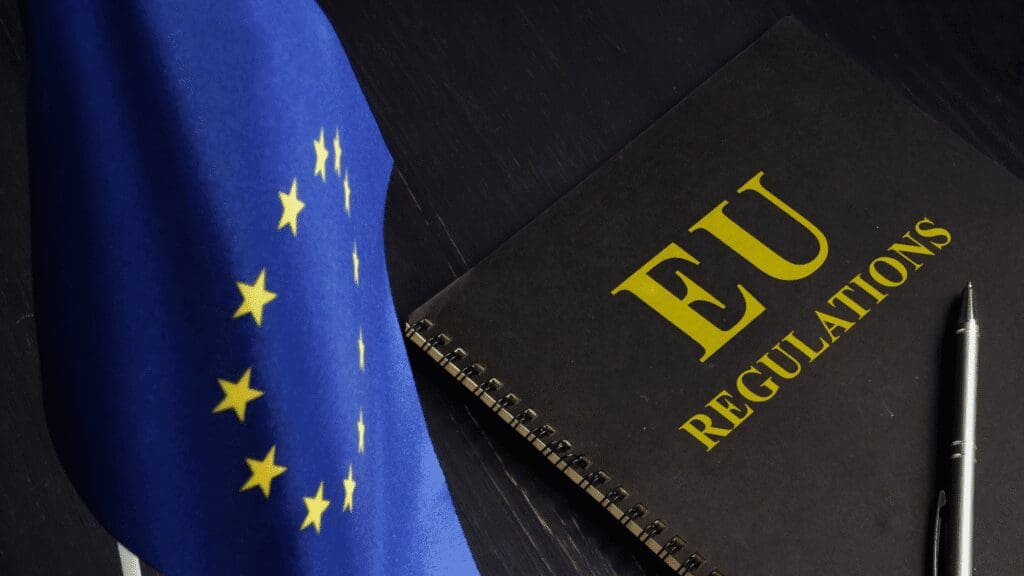 Close-up of an EU flag next to a black spiral notebook labeled "EU Regulations" with a pen lying beside it, symbolizing European Union legislative and regulatory processes.
